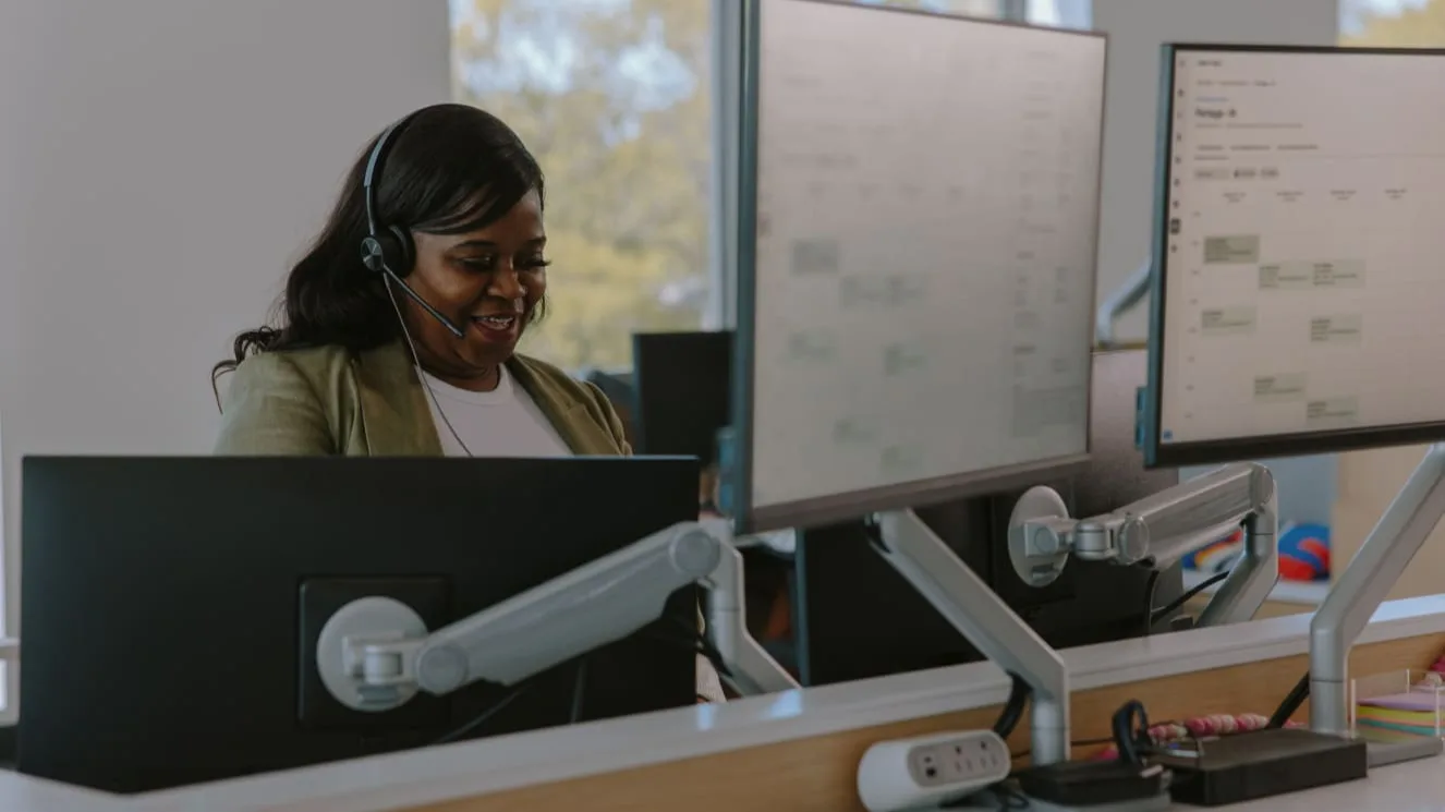Customer service representative wearing headset smiles while working at desk with multiple monitor setup in bright office.