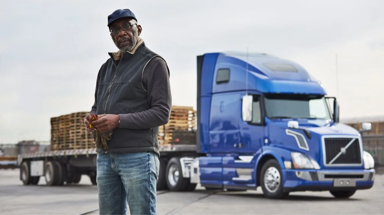 Person wearing cap and vest standing in front of blue semi truck with flatbed trailer loaded with wooden pallets.