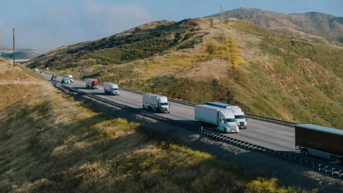Semi trucks traveling on highway winding through golden hills under blue sky, with communication towers on distant peaks.