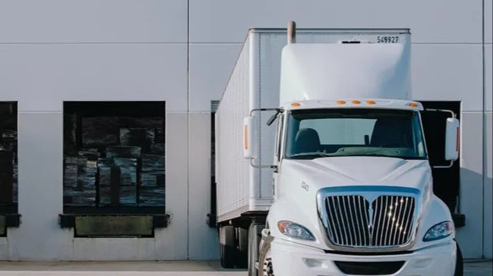 White delivery truck parked at loading dock with warehouse doors visible against a light-colored building.