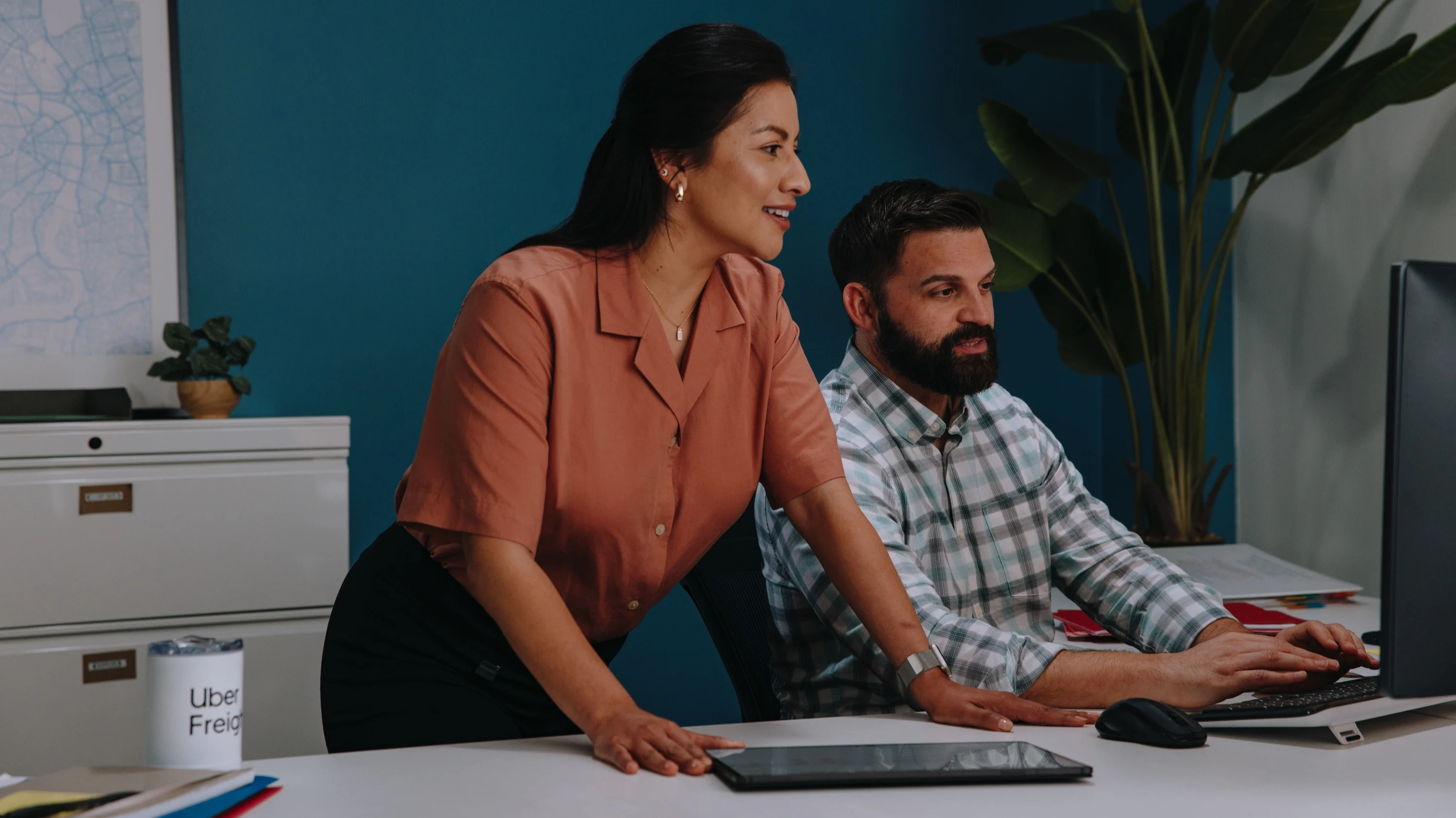 Two professionals working together at a desk with computer and tablet against a teal wall with indoor plants.