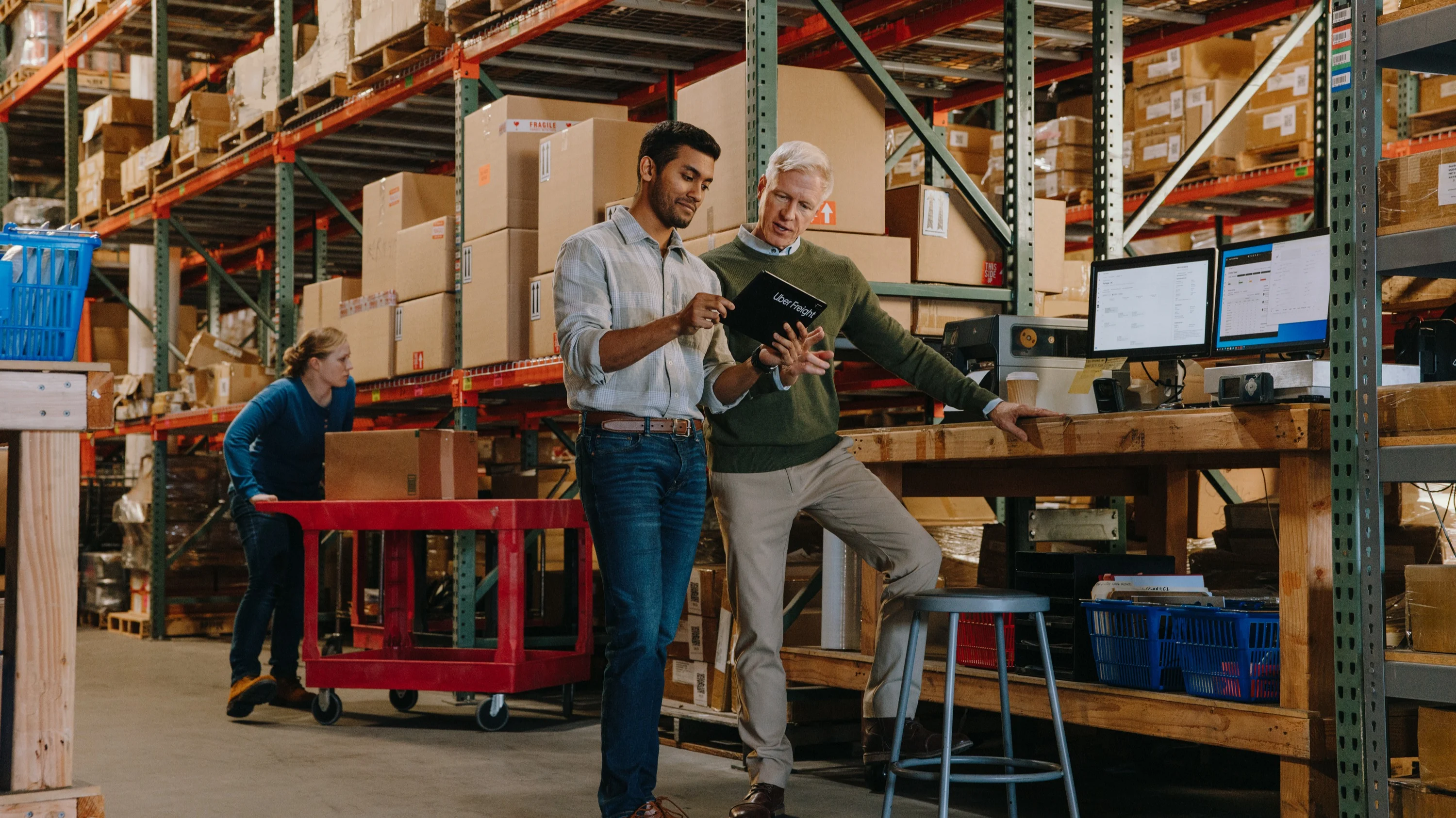 Person in green sweater working at computer terminal in warehouse with shelves of boxed inventory in background.